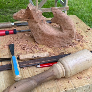 Ein aus Holz gehauener Drache steht auf einer Werkbank mit Werkzeugen zum Holzbildhauen, wie Klüpfel und Stecheisen