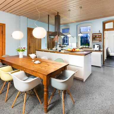 Wohnküche Freistehende Küchenzeile auf einem grauen Mosaikboden, ein Holztisch mit Stühlen im Vordergrund und einem Holzofen