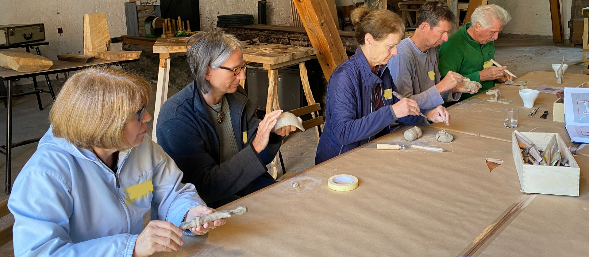 5 Menschen sitzen an einem langen Tisch und formen mit den Händen Modelle aus Ton