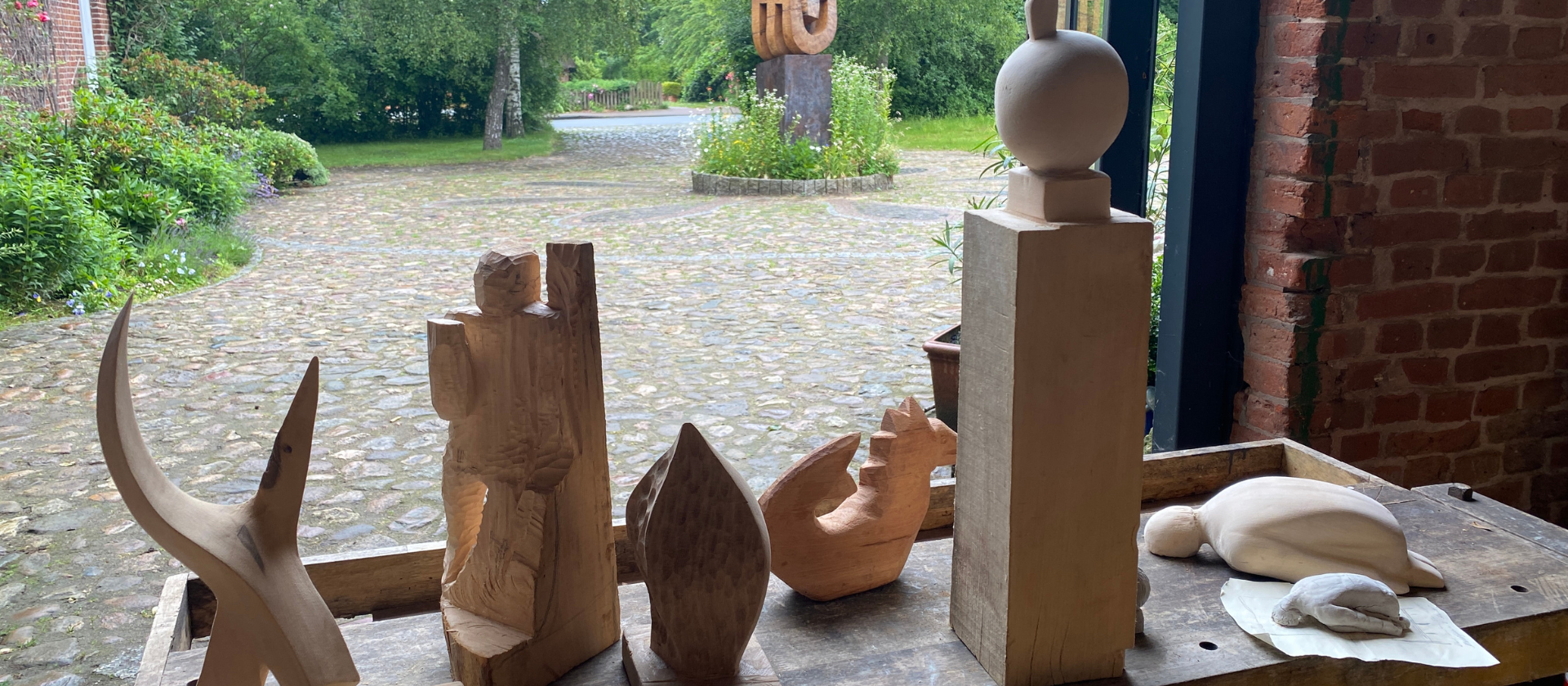 Einige verschiedene Holzskulpturen, die in einem Workshop im Werkgut gefertigt wurden. sie stehen auf einer Werkbank, im Hintergrund ist ein Hof mit einer großen Skulptur zu sehen.