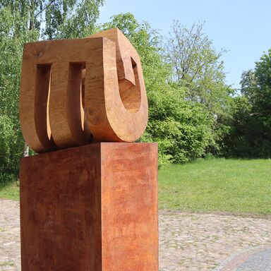 Skulptur "Niemandsblau" Holzskulptur von Yves Rasch auf kupferfarbenem Sockel in der Einfahrt vor Rasen und Bäumen