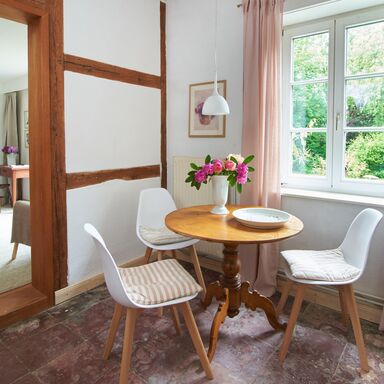 Essbereich in der Künstlerwohnung drei Stühle an einem runden Tisch mit Blumen in einer Vase vor einem Fenster mit rosa Gardinen