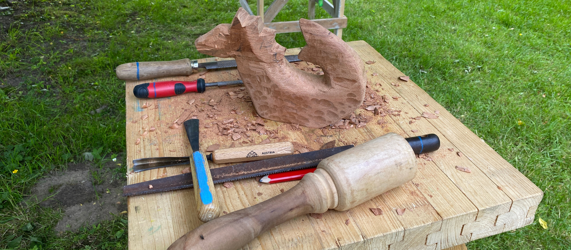 Ein aus Holz gehauener Drache steht auf einer Werkbank mit Werkzeugen zum Holzbildhauen, wie Klüpfel und Stecheisen