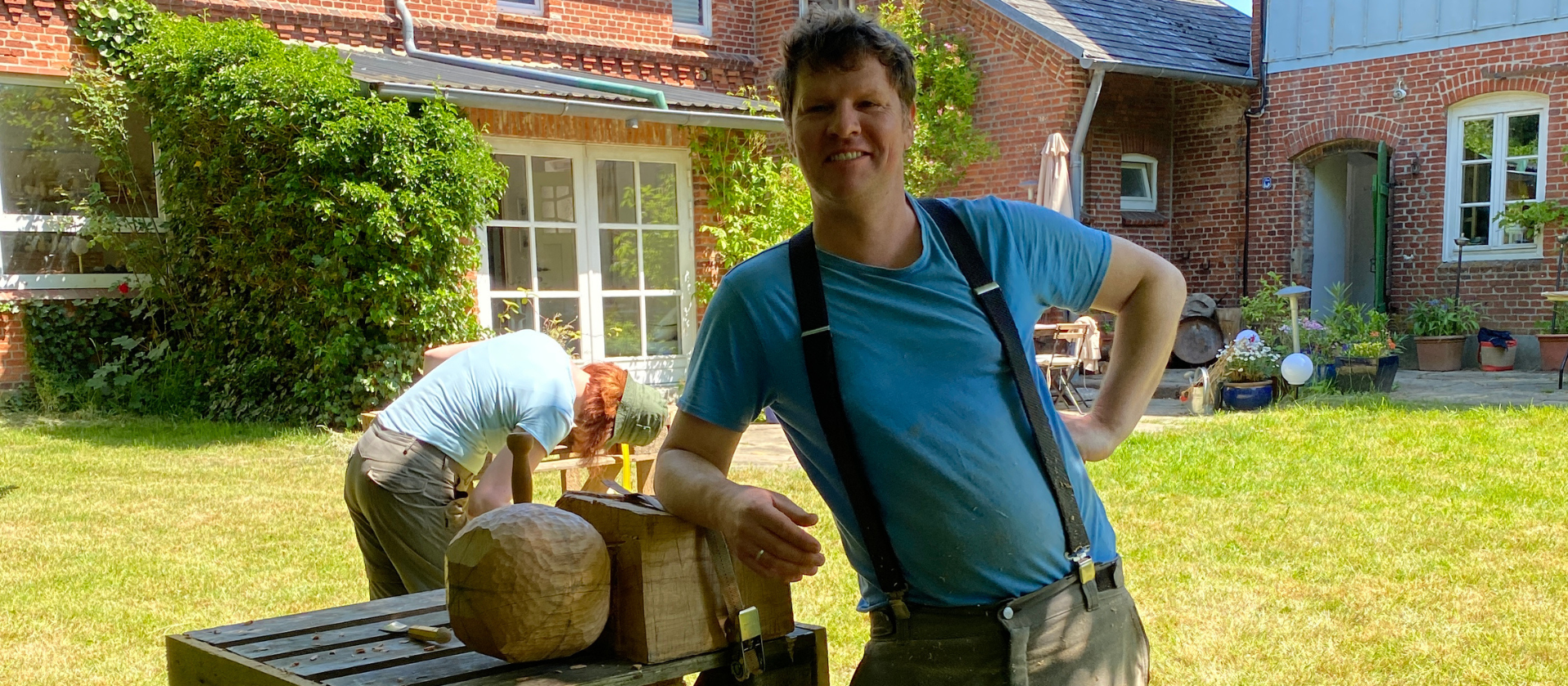 Holzbildhauer Yves Rasch locker mit einem Arm auf einer unfertigen Holzskulptur lehnen im Garten des Werkgut