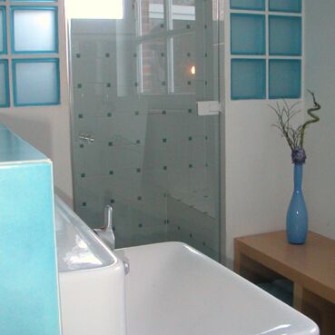 weißes Waschbecken im Vordergrund, im Hintergrund eine gläserne Duschtür und eine Sitzbank aus Holz mit blauer Blumenvase