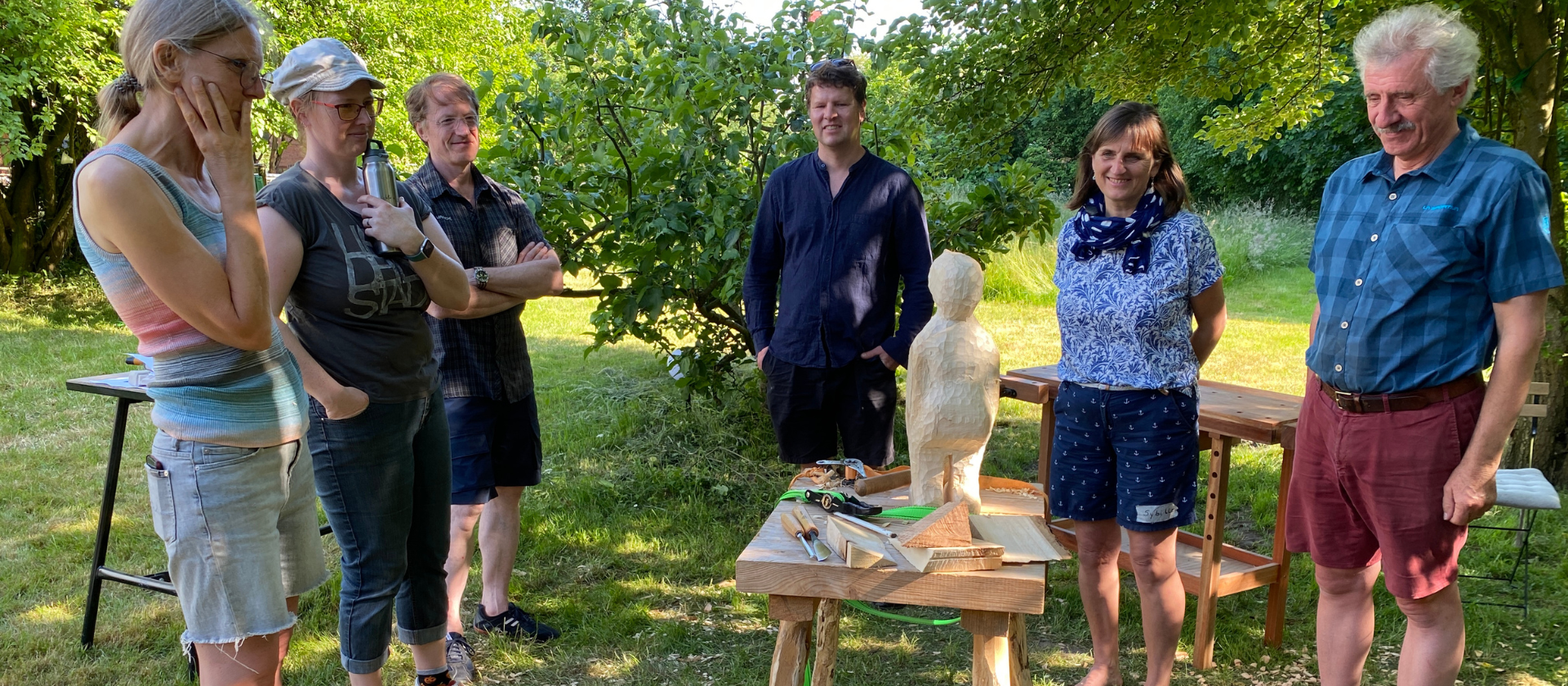 Die Gruppe des Workshops steht um eine Holzbildhauen Skulptur herum und betrachtet das Werk