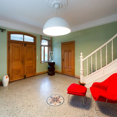 Eingangshalle Eingangsbereich des Werkguts mit grauem Mosaikboden, einer weißen Treppe, die nach oben führt und einem leuchten roten Sessel mit Hocker.