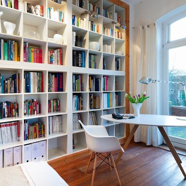 Ein Stuhl neben einem Tisch und einer Leseleuchte vor einem Fenster mit Blick ins Grüne, dahinter ein deckenhohes gefülltes Bücherregal.