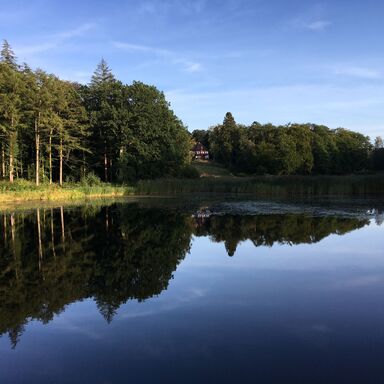 See an einem klaren Tag, der Himmel spiegelt sich in der glatten Wasseroberfläche. Am Ende des Sees, sieht man Waldhütten