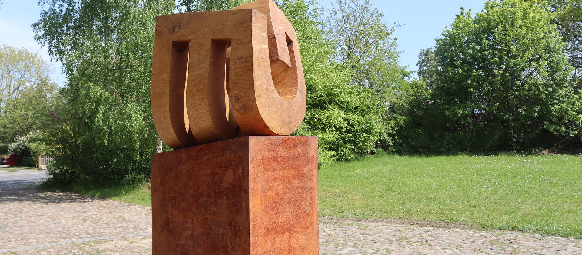 Skulptur "Niemandsblau" Holzskulptur von Yves Rasch auf kupferfarbenem Sockel in der Einfahrt vor Rasen und Bäumen