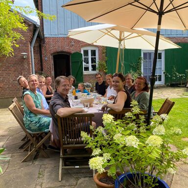 Eine große Gruppe Workshopteilnehmer sitzt am Tisch auf der Terrasse unter einem großen Sonnenschirm. Alle blicken fröhlich in die Kamera