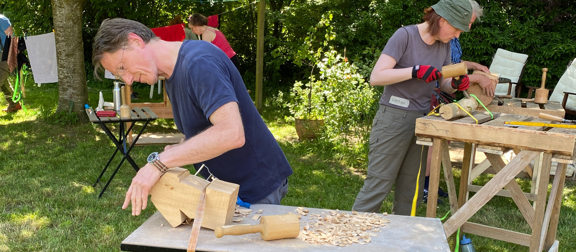 Links arbeitet ein Mann mit Brille in blauem T-Shirt an einem Stück Holz, das auf einer Werkbank festgespannt ist. Ein Klüpfel liegt neben ihm. Rechts im Hintergrund arbeitet eine Frau mit Stecheisen und Klüpfel an ihrer Holzskulptur.