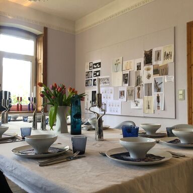 Elegant gedeckter Tisch mit Porzellan von Mikaela Dörfel, in der Vase sind Tulpen, an der Wand hängt eine Tafel mit Entwürfen