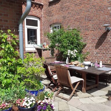 Terrasse Auf der steinernen Terrasse stehen ein gedeckter Gartentisch und Stühle, im Vordergrund einige Blumentöpfe mit Blumen und Pflanzen