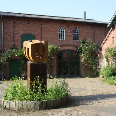 Der Hof Skulptur mit Bepflanzung am Sockel auf dem Hof vor dem Tor zur Diele