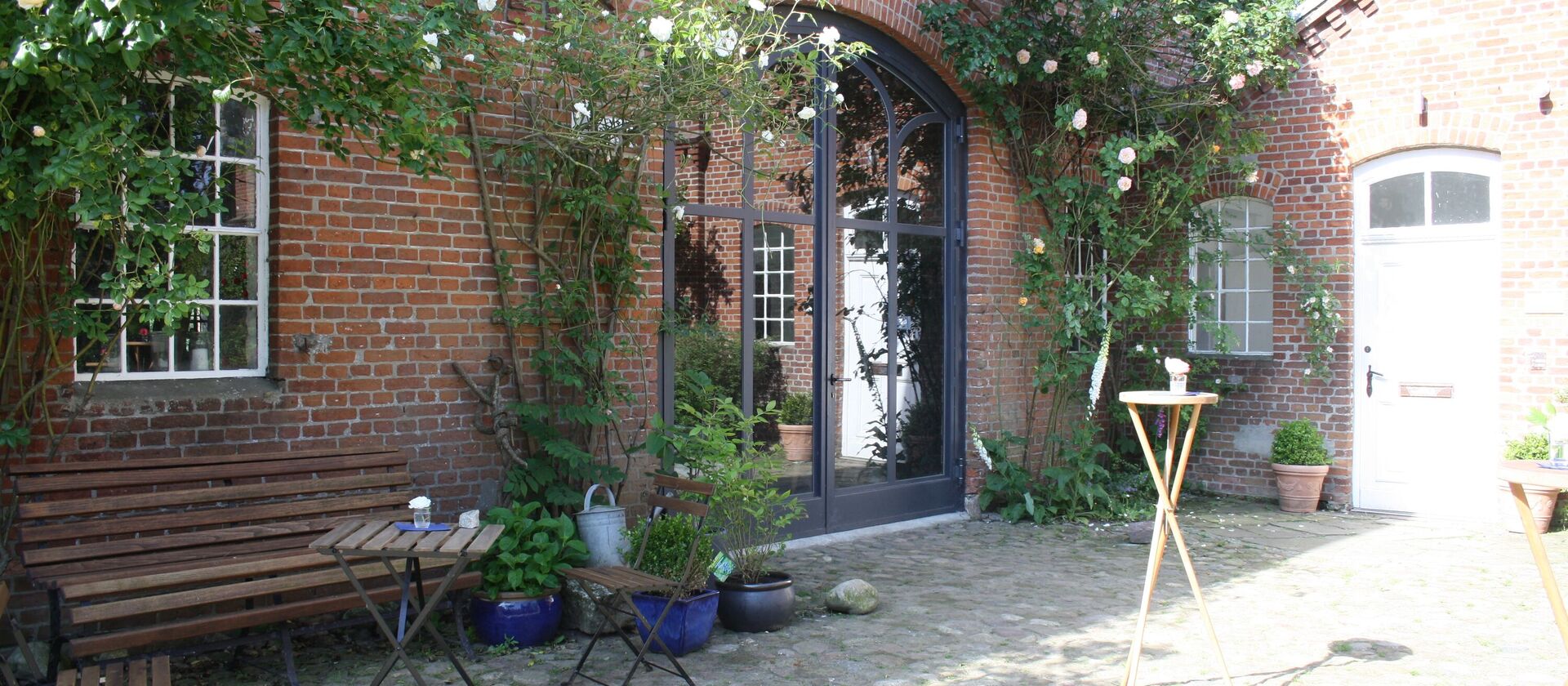 Im Vordergrund links eine Holzbank an der Wand mit zwei Stühlen und einem Tisch und rechts ein paar Stehtische, im Hintergrund das geschlossene Tor zur Diele des Werkguts