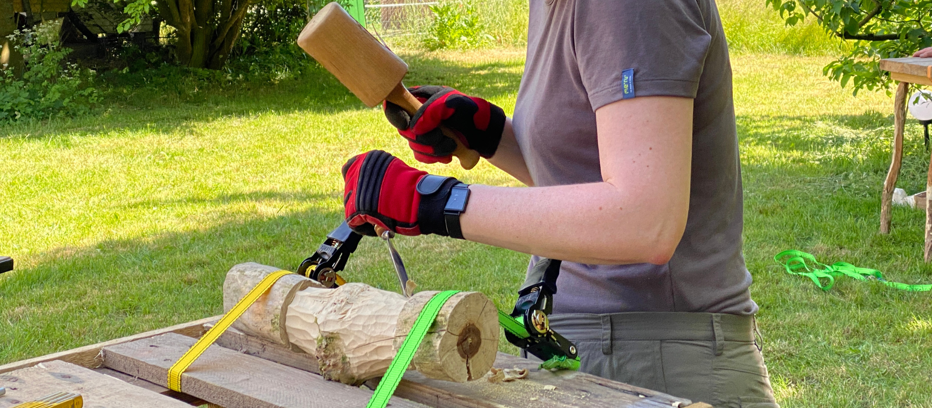 Eine Person arbeitet mit Klüpfel und Stecheisen an einem Stück Holz, das mit zwei Spanngurten an einer Werkbank befestigt ist.
