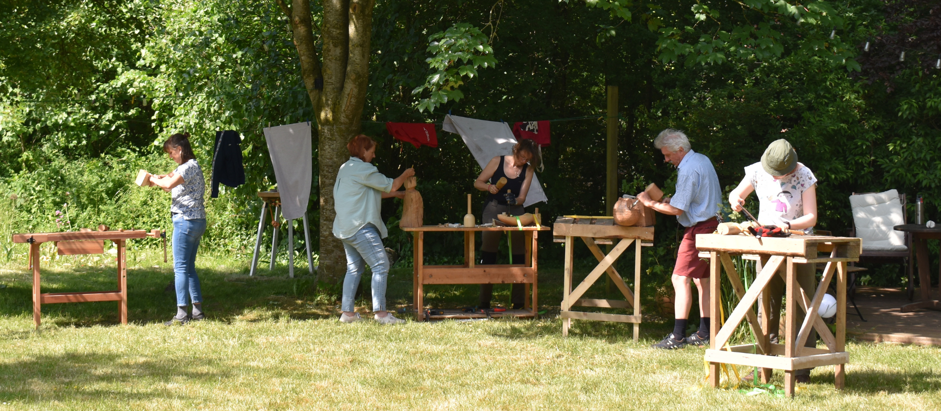 Im Garten stehen vier Werkbänke und fünf Menschen arbeiten an ihren Holzskulpturen.