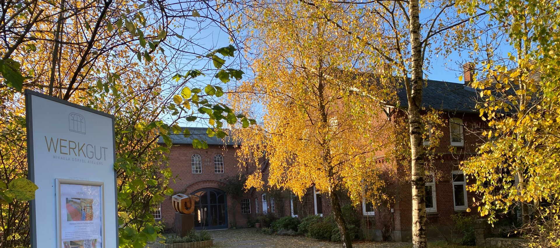 Hof im Herbst Links eine Tafel mit der Aufschrift Werkgut und einem Plakat und Sicht auf die Einfahrt des Werkguts mit herbstlichen Bäumen