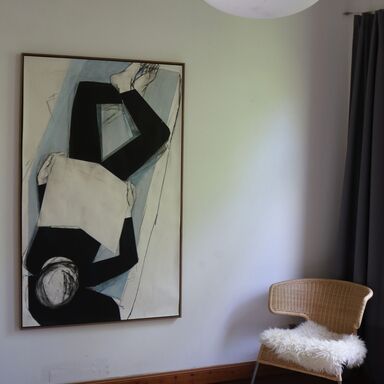 An der Wand hängt ein halbabstraktes Gemälde mit einer liegenden Person, die ein Buch liest. Neben dem Bild steht ein Rattanstuhl mit Schaffell auf der Sitzfläche