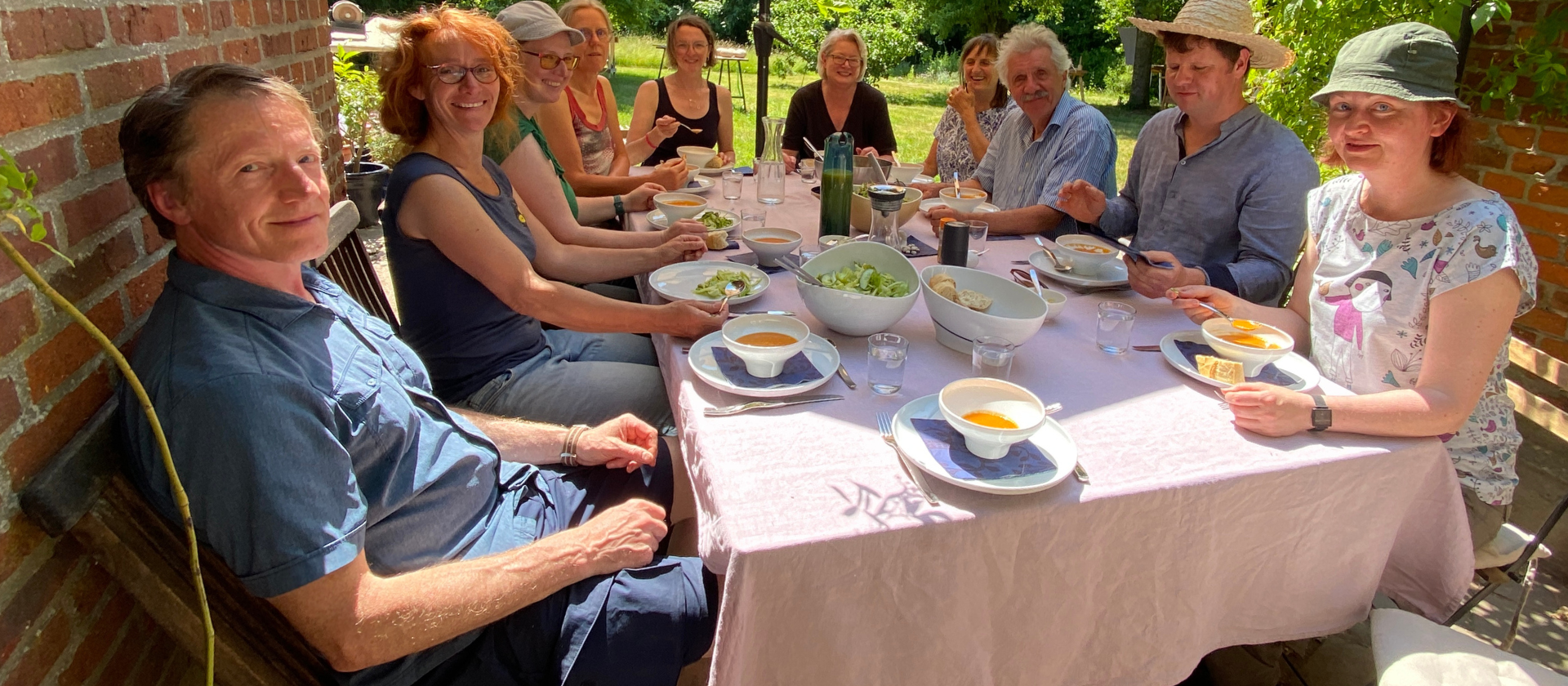 Einige Menschen die an einem sonnigen Tag an einem Tisch sitzen und Suppe essen. Alle sind fröhlich und lächeln in die Kamera.