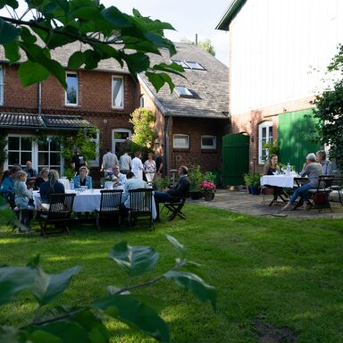 Gartenparty Blick auf den Garten mit am Tisch sitzenden Menschen, die feiern. Im Hintergrund das Haupthaus, im Vordergrund Blätter